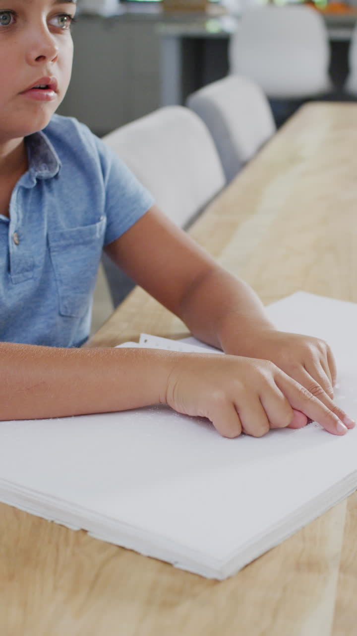 Vertical video of caucasian boy sitting at table and reading braille, slow motion