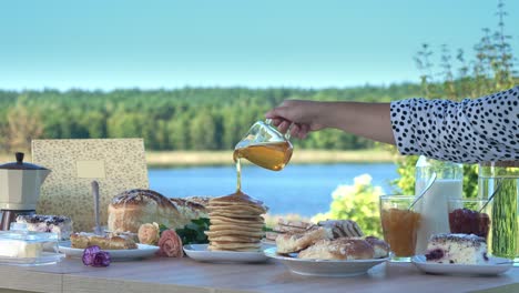 woman's hand pouring maple syrup from a glass cup to a stack of pancakes in a plate - breakfast outdoor - static shot