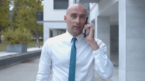 vista frontal de un hombre de negocios confiado en camisa blanca, hablando por teléfono móvil mientras camina cerca del edificio de oficinas afuera