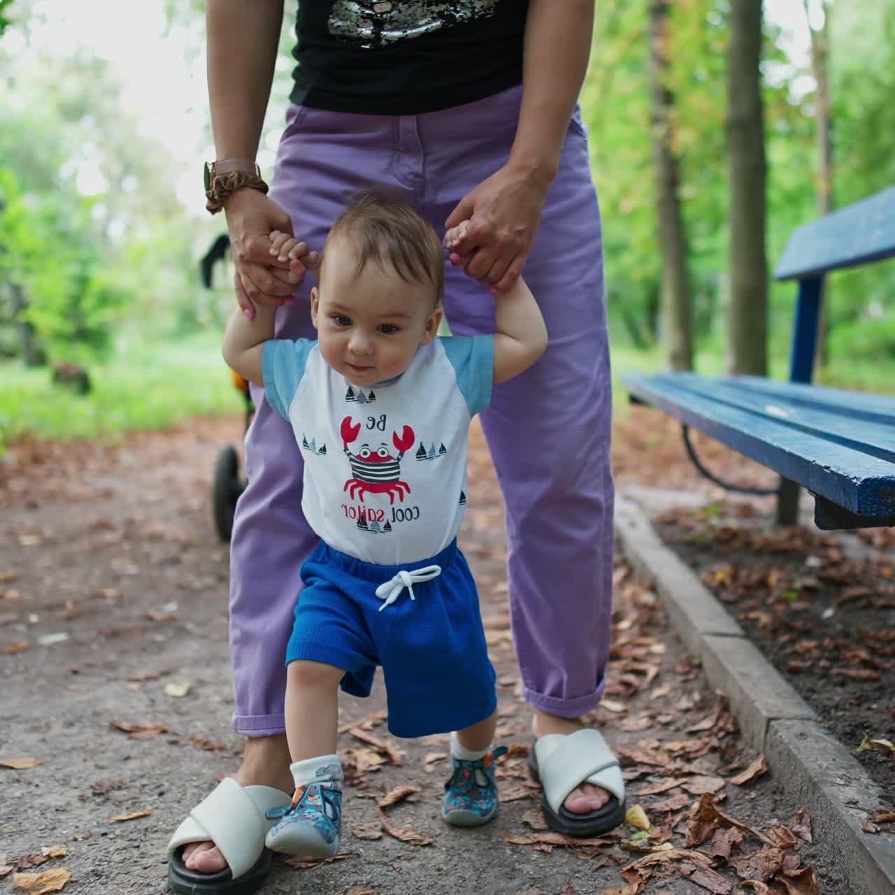 Lovely cute Caucasian boy walks supported by his mom. Little kid stepping with interest and adorable smile. Green park backdrop