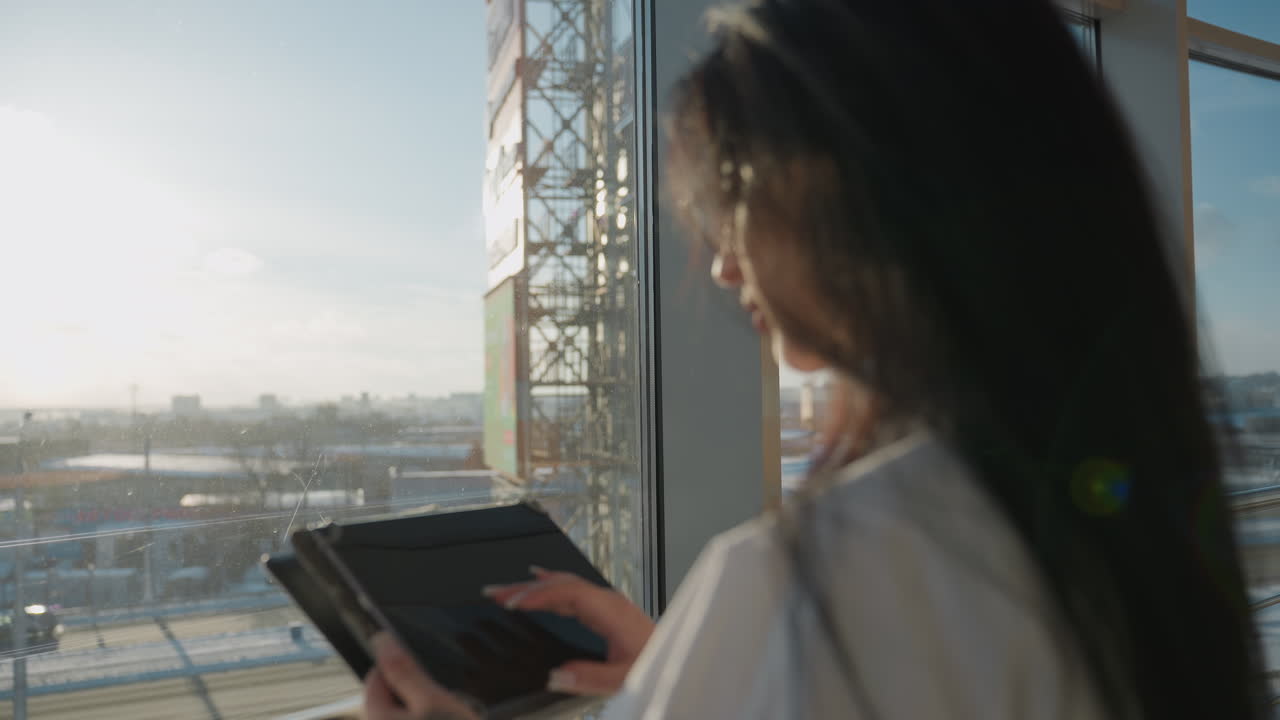 Professional woman adjusts glasses while holding tablet, looking through glass window at vehicles moving along snowy road, with tall urban structure visible under clear blue sky and bright sunlight
