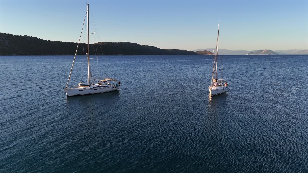 Greece,Kalamos Island,Aerial view forward towards Kastos island passing in between the two white sailboats anchored. Calm seas and crystal clear waters on a sunny day