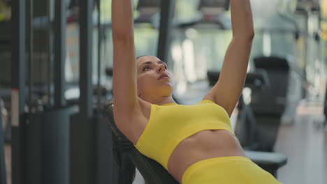 Determinada-Atleta-Hispana-Haciendo-Press-De-Banca-Con-Pesas-En-El-Gimnasio.-Una-Joven-En-Forma-Haciendo-Un-Press-De-Banca-Con-Mancuernas-En-Un-Pequeño-Gimnasio.-Culturista-Femenina-Musculosa-Tumbada-En-El-Banco-Y-Haciendo-Pesas