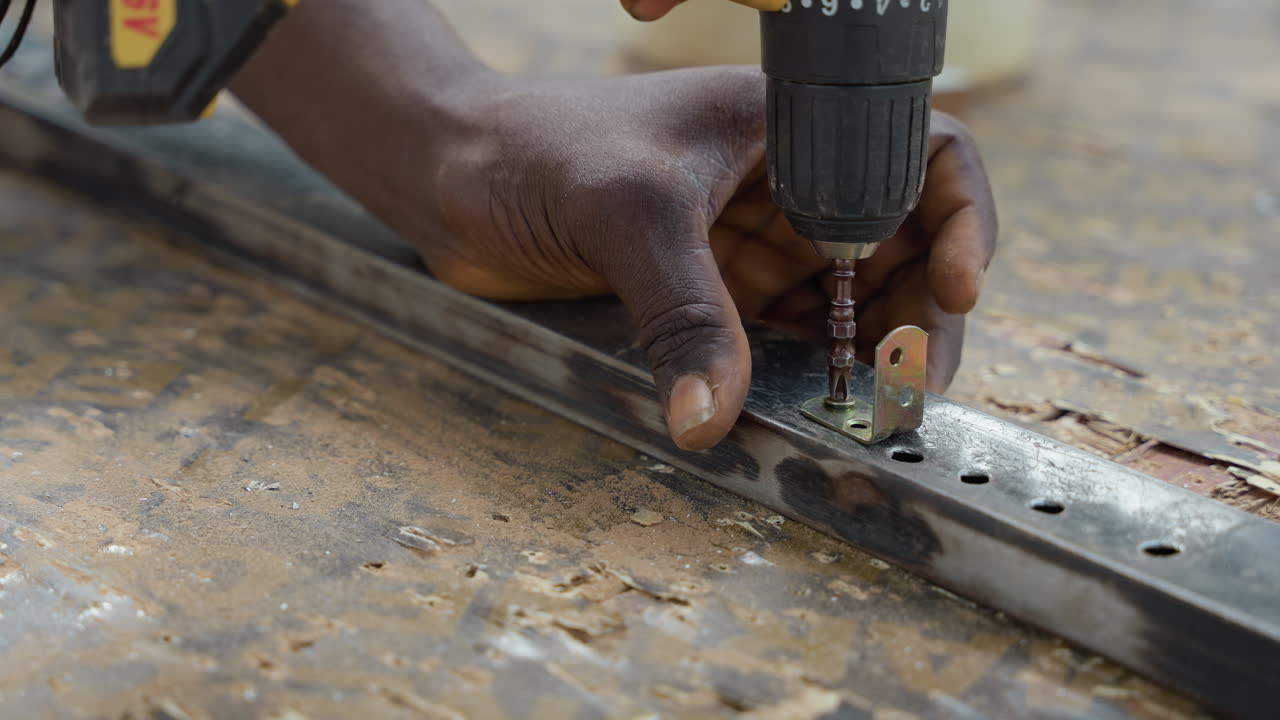 Primo piano di una mano che usa un trapano per fissare una vite in un binario metallico su un banco da lavoro usurato, allineamento di una piccola staffa, trucioli di metallo e polvere, presa manuale ferma, foratura precisa e assemblaggio di mobili, concentrato