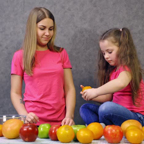 mother and daughter working together in the kitchen. healthy organic fruit on the table. beautiful woman and little girl peeling fruits.