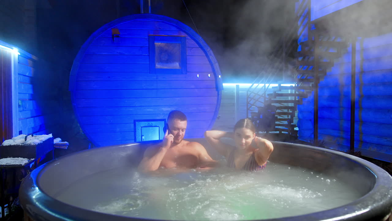 Relaxing couple sit in hot tub outdoors in winter. Man speaks on the phone and woman touches her hair.
