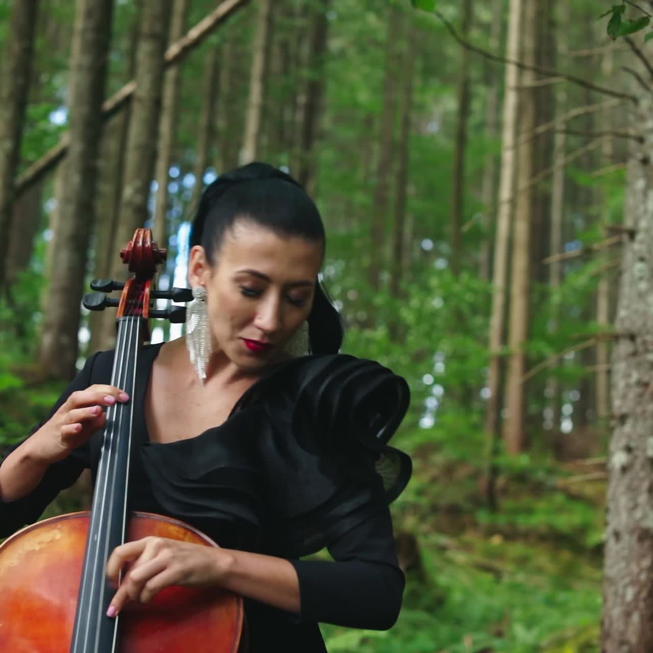 Beautiful cellist in the forest. Luxury woman in black dress playing the cello on green nature background. Attractive female musician performing music outdoors.