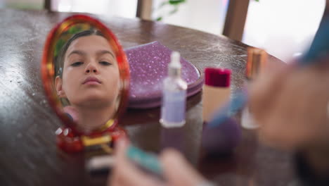 close-up view of girl applying makeup on her face while looking into mirror reflection, blurred cosmetic items like bottles, powders, and a pouch are visible on the table