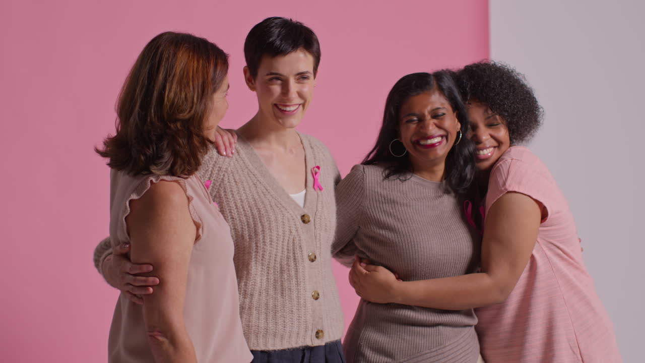 retrato de estudio de un grupo multirracial de mujeres de diferentes edades que llevan cintas rosadas de concienciación sobre el cáncer de mama abrazándose contra un fondo rosa 2