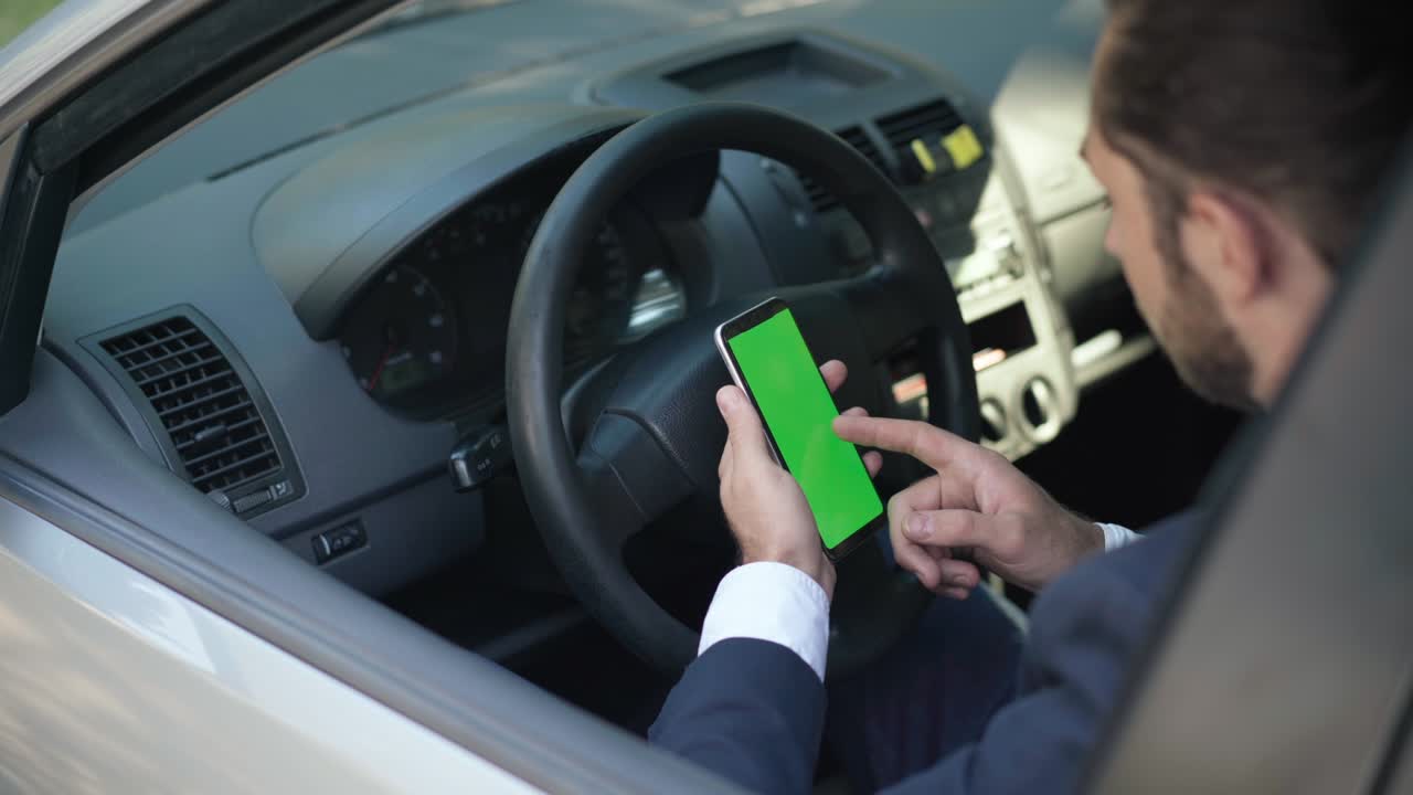 Young Caucasian man swiping chromakey smartphone zoom in screen sitting on driver's seat waiting. Confident male driver surfing Internet using social media in car. Modern technologies and lifestyle.