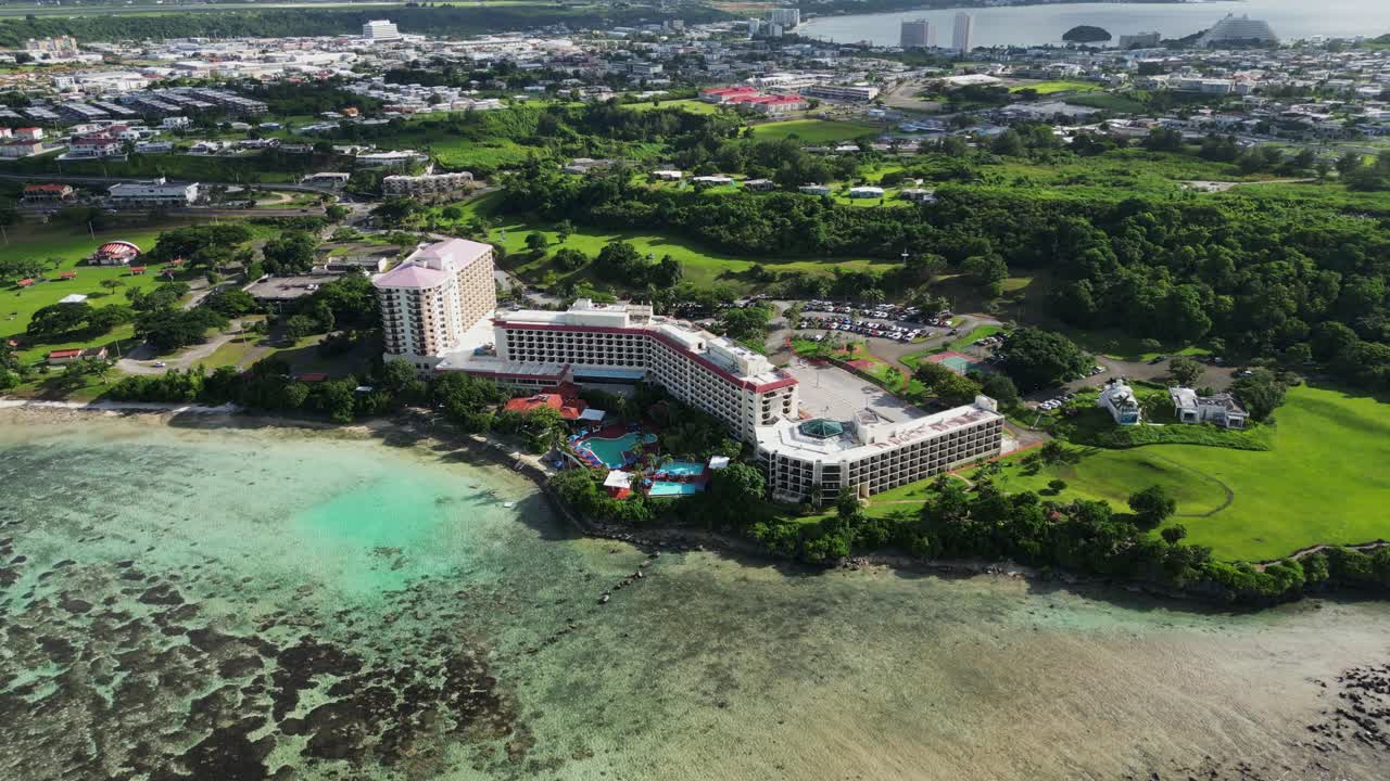 Picturesque view of coastal hotel resort with lush greenery facing shallow ocean reef at tropical island Guam, USA - aerial orbit