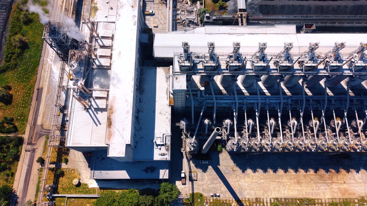 Vista aerea dall'alto di torri e tubazioni di un impianto industriale di lavorazione. File di recipienti di lavorazione in metallo e tubazioni collegate creano ombre geometriche sul terreno in cemento.