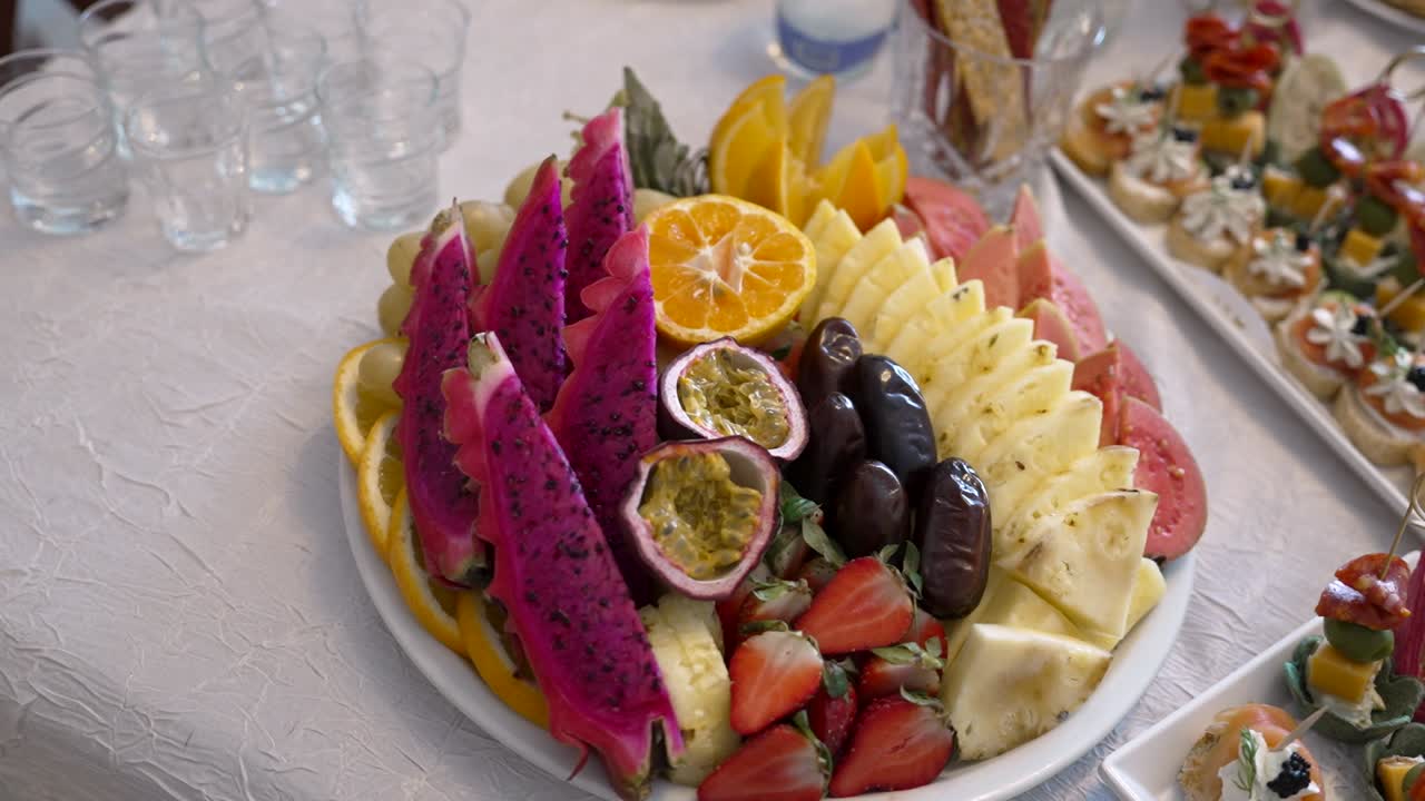 Fargerike frukter vises på et stort fat under en sammenkomst. Arrangementet inkluderer druer, jordbær, ananas og eksotiske frukter som dragefrukt og pasjonsfrukt.