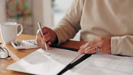 a man sits at a table, writing on a piece of paper with a pen and calculator. he is wearing glasses and has a cup of coffee on the table beside him.