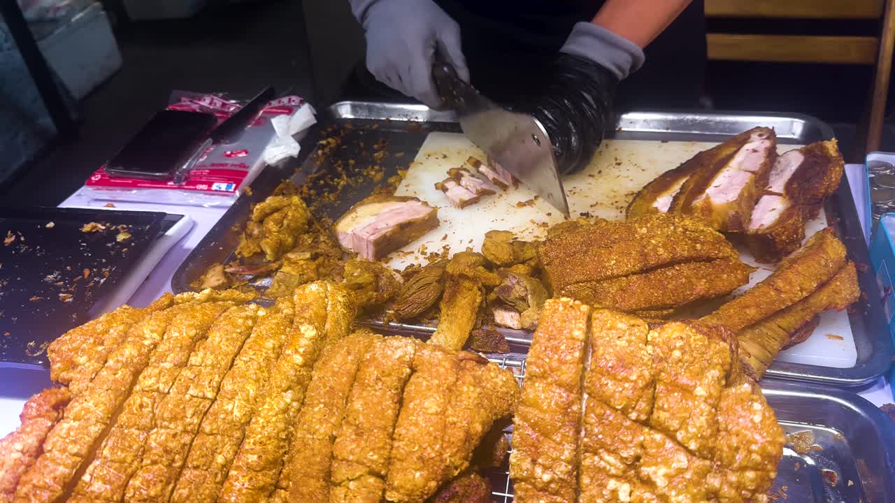 A vendor slices crispy pork at a bustling Bangkok market. Bright lighting highlights the golden texture, capturing the vibrant street food scene