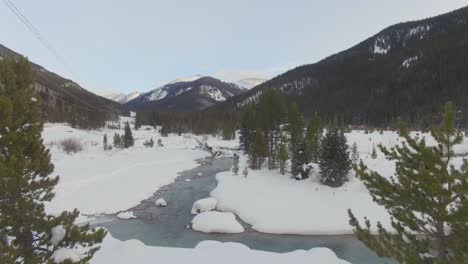 slow fly through small trees over winding river in snow covered mountain valley