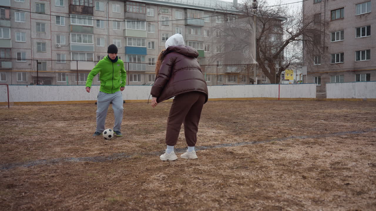 진흙 투성이의 안뜰 경기장에서 흰색 남성과 흰색 노년 여성 간의 친선 축구 결투, 패스와 태클 주고받기, 아파트 블록 배경, 캐주얼 재킷과 스니커즈, 장난기 넘치는 경쟁