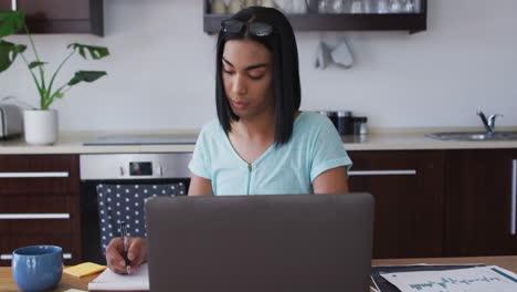 persona de género mixto fluida sentada en un escritorio tomando notas trabajando desde casa usando una computadora portátil