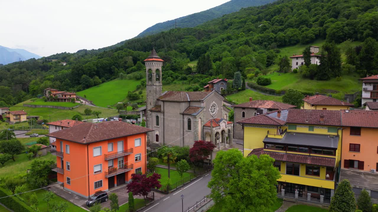 Drone shot of a stone church surrounded by colorful homes and green hills in the Alpine foothills. Shot at Villa di Tirano, Sondrio, Italy (Italia)