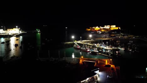 Nächtliche-Schwenkaufnahmen-über-Den-Hafen-Von-St.-Peter-Port,-Guernsey,-Mit-Beleuchtetem-Castle-Cornet-Im-Hintergrund-Und-Detaillierter-Ansicht-Der-Festgemachten-Fischerboote