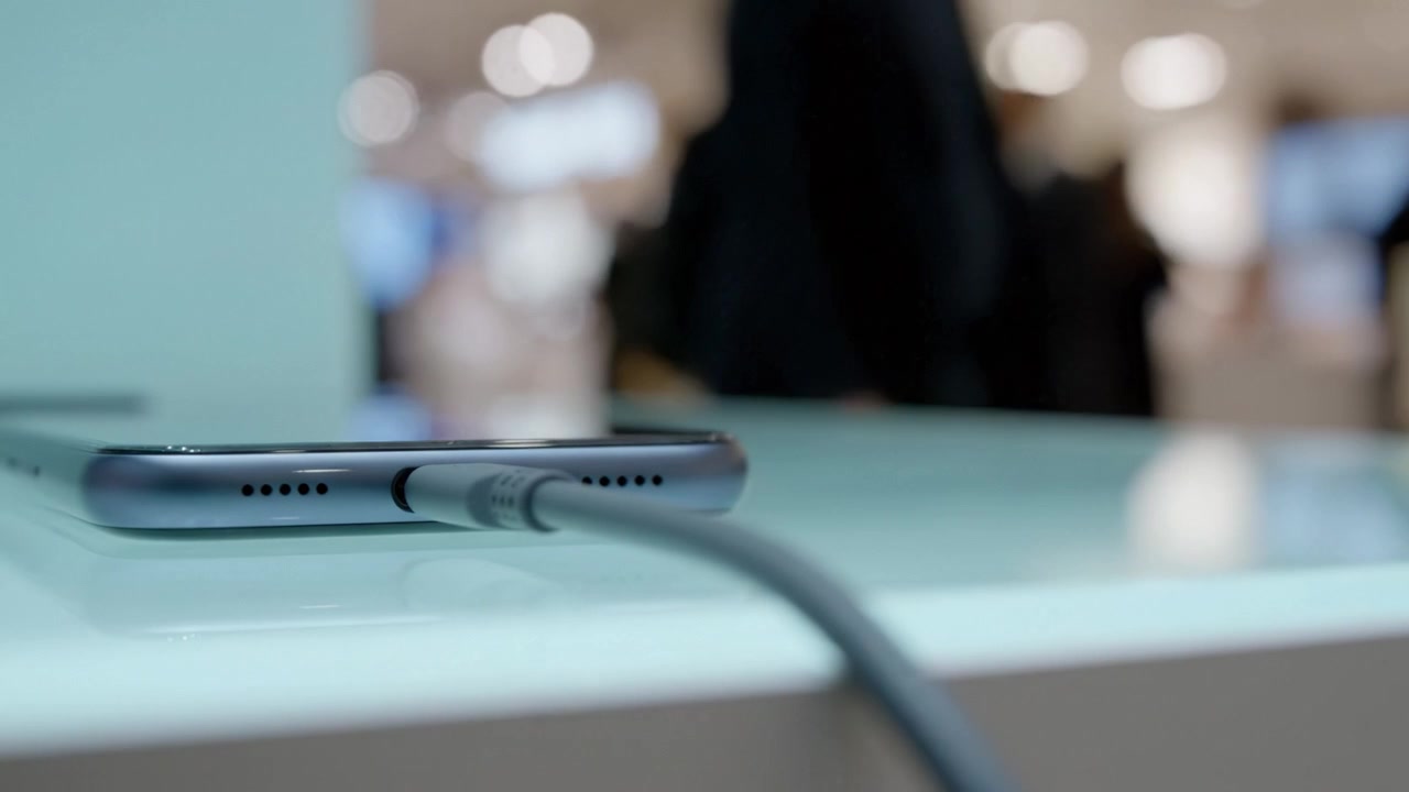 Una Vista Ravvicinata di uno Smartphone in Carica con un Cavo su un Display Moderno, Circondato da un'Atmosfera Luminosa Piena di Attività Sfocata sullo Sfondo