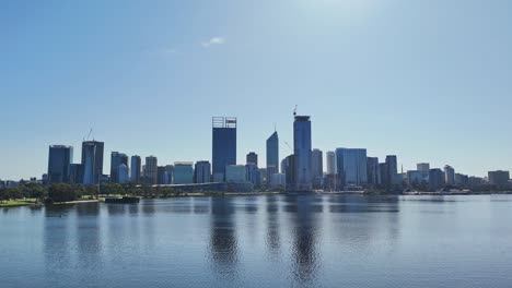 Drone-flying-along-swan-river-in-Perth,-Western-Australia-on-sunny-day-with-silhouette-of-city-skyline-in-the-background