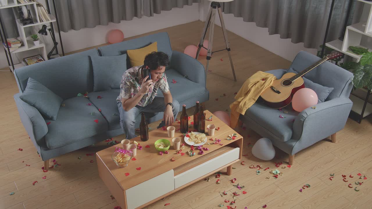 vista superior de un hombre asiático borracho sentado en un sofá y hablando en su teléfono inteligente después de tener una fiesta en casa