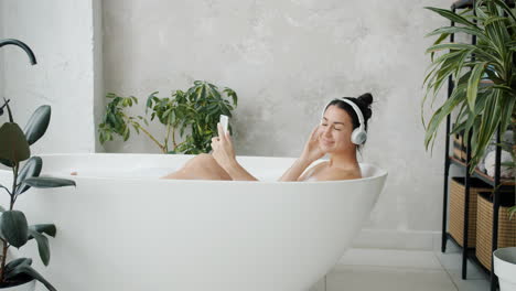 woman relaxing in a bathtub with headphones and a smartphone