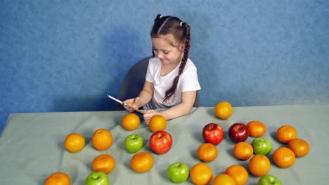 happy child with many organic fruit in the kitchen. little girl playing on digital tablet and counts fresh fruits on the table.