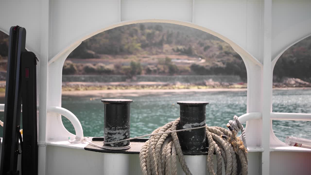 A shoreline moves through the frame of a ferry port and an anchor rope.