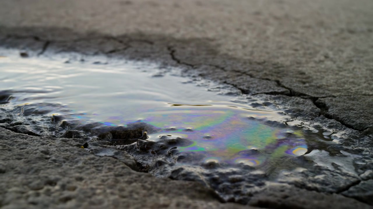 Close-up dari permukaan tanah yang memperlihatkan genangan air dangkal di aspal retak yang terkikis, dengan lapisan minyak tipis menciptakan warna pelangi yang berkilauan di atas air yang memantul, di mana riak dan guratan halus terlihat