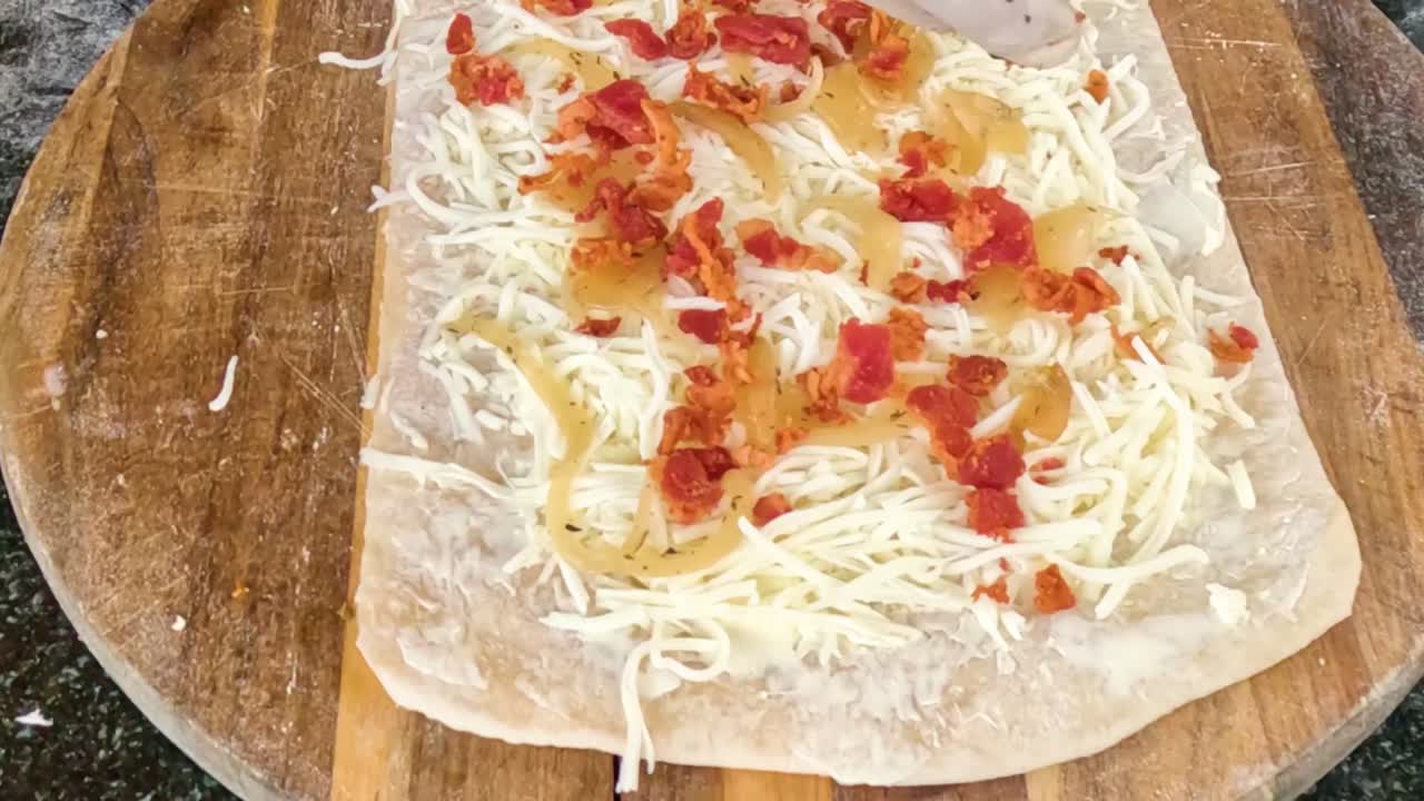 A gloved hand adds cheese and diced tomatoes onto flatbread on a wooden board.