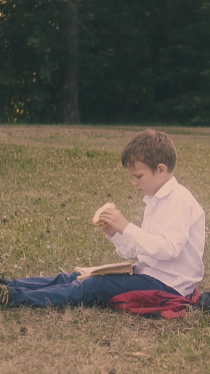 concentrated junior pupil in white shirt eats hamburger sitting on grass near reading classmates close view slow motion
