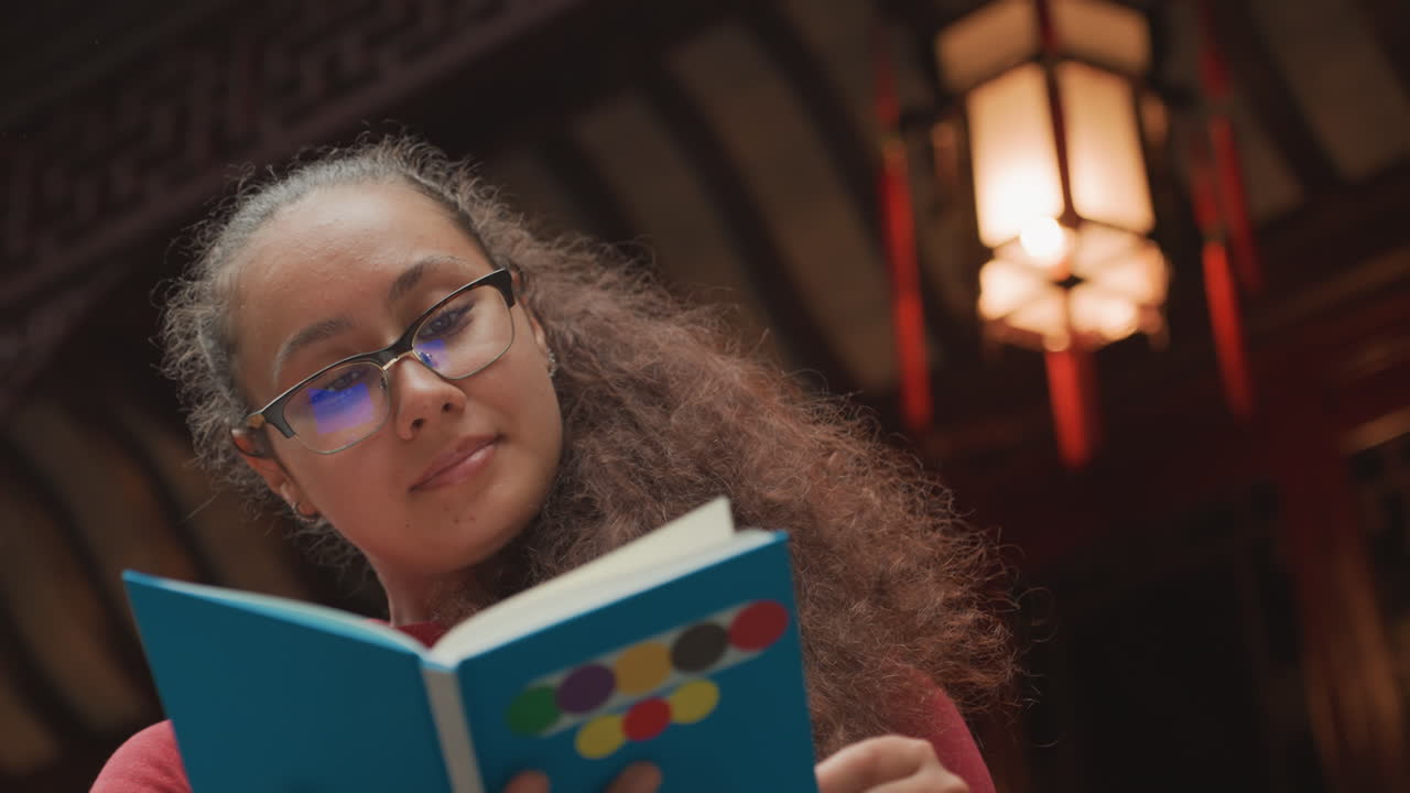 Giovane donna che legge un libro blu in un accogliente interno illuminato da lanterne, che indossa occhiali e ha i capelli ricci, espressione concentrata mentre sfoglia le pagine, travi di legno calde e luce ambientale soffusa creano un'atmosfera tranquilla e pensierosa