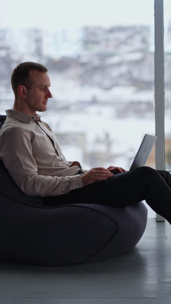 Middle-aged male works on laptop sitting in bean bag chair. Man distracts and looks aside, putting hand to his forehead. Cityscape in blur at backdrop. Vertical video