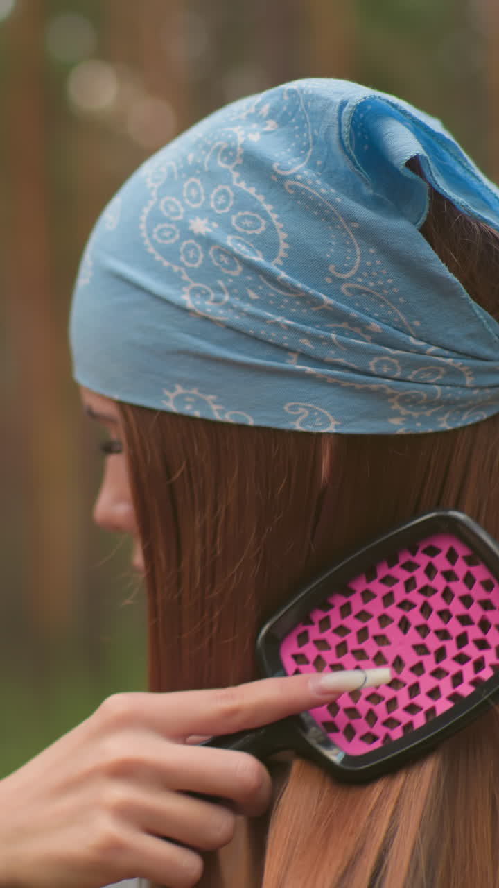 primer plano de una mujer cepillándose el cabello mientras está en un entorno forestal sereno, con un pañuelo azul atado alrededor de su cabeza, en el fondo borroso, otra mujer se centra en abrir su termo