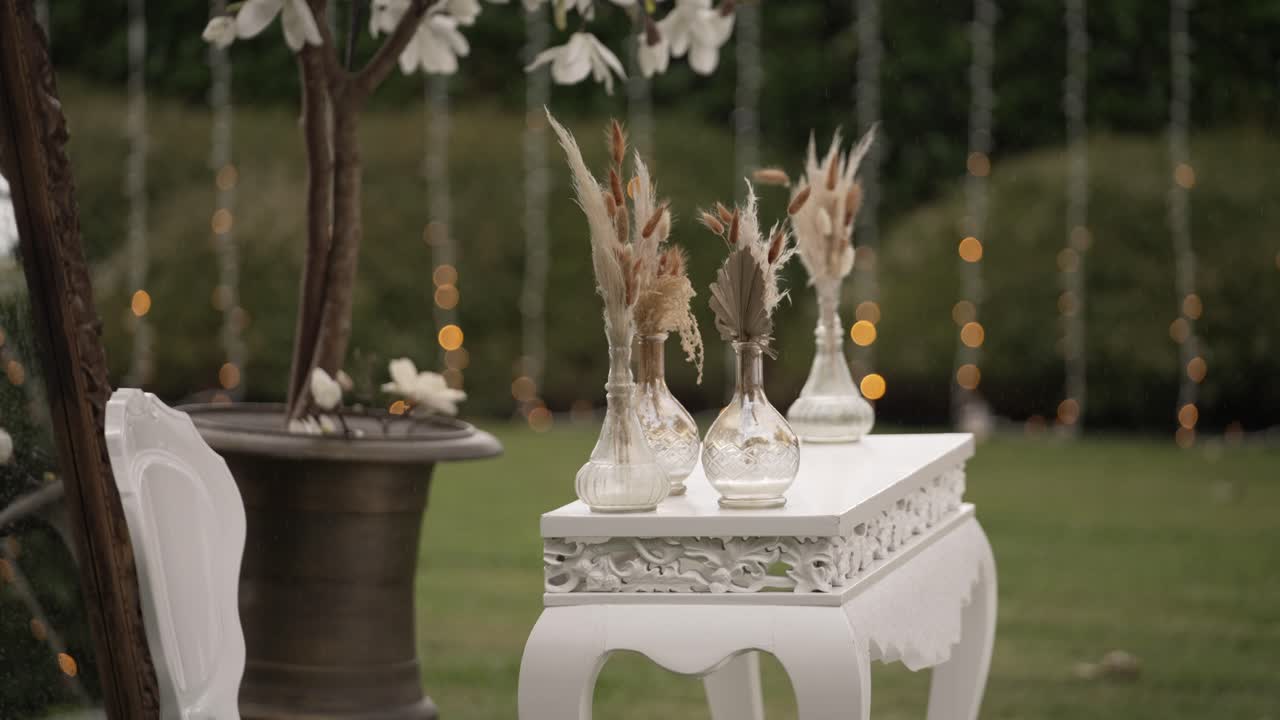 Decorative table with dried floral arrangements in vases set outdoors, lights in background