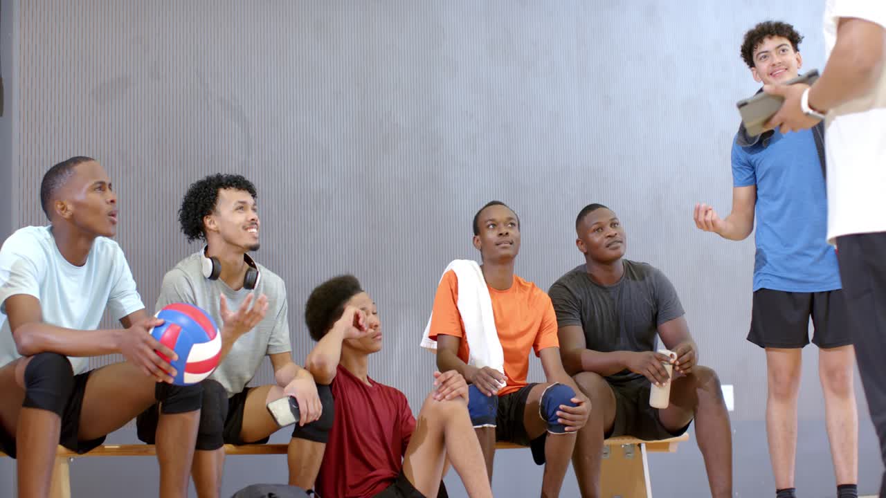 Diverse Athleten und ihr Trainer sitzen auf einer Fitnessbank und nutzen ein Tablet, während ein Athlet die Hand hebt, um zu pausieren und zu antworten.