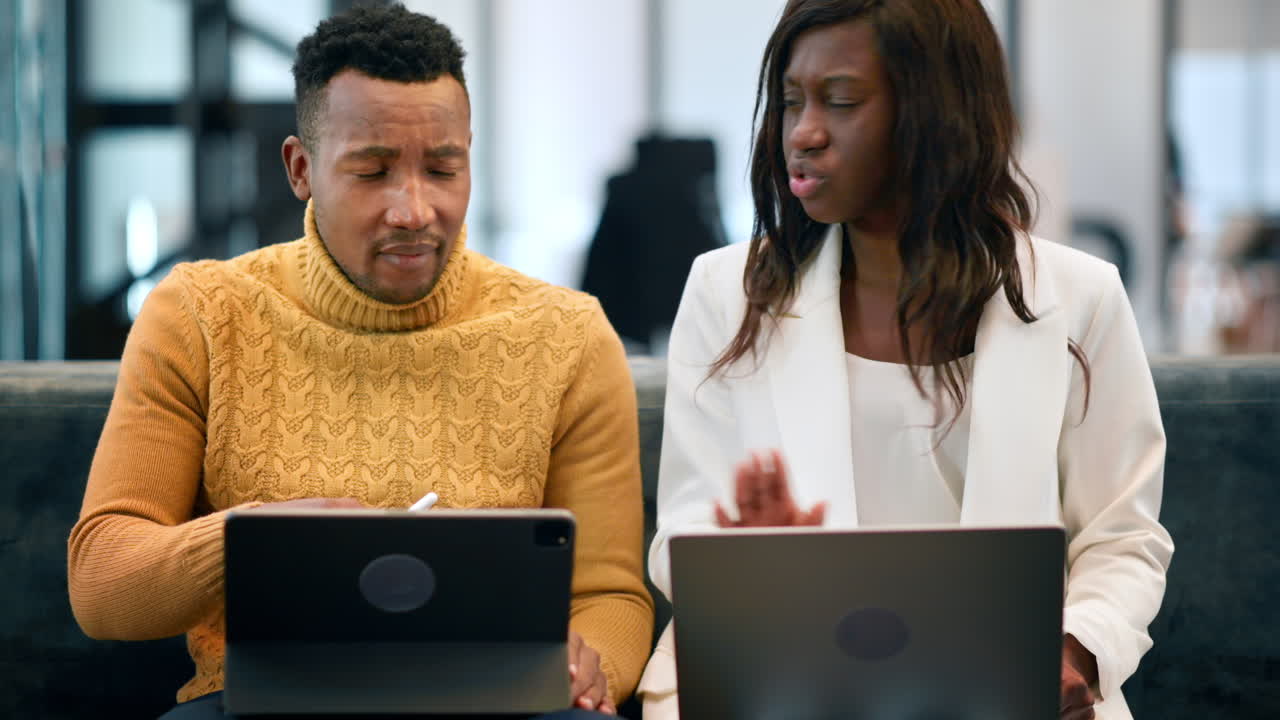 Black man and woman working using tablet and laptop in an office, discussing business affairs while sitting on a sofa