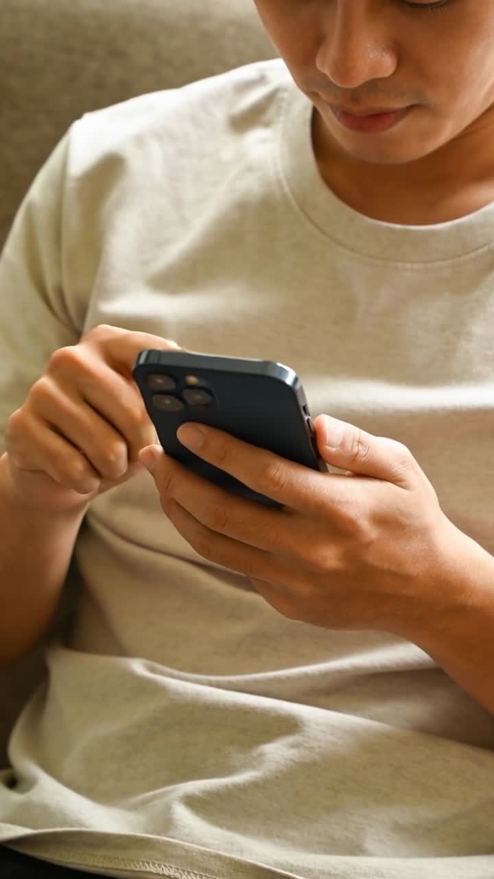 Vertikální video: Asijský muž držící smartphone, ťukající a kontrolující zprávy na pohovce, v béžovém tričku