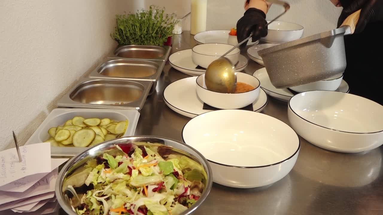 Flere skåle varm suppe serveret med ristet brød på tallerkener i et restaurantkøkken, med fritter, salat og syltede grøntsager synlige, mens en handske-klædt hånd anretter retterne