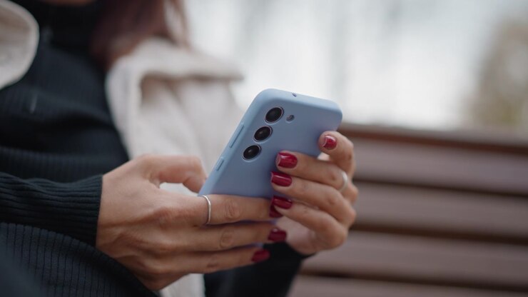 Vista ravvicinata di mani di adulto maturo che tengono uno smartphone azzurro pallido su una panchina di legno del parco, con i pollici che toccano e scorrono con manicure rosso scuro, mostrando un uso mobile casuale e concentrato all'aperto alla luce soffusa del giorno