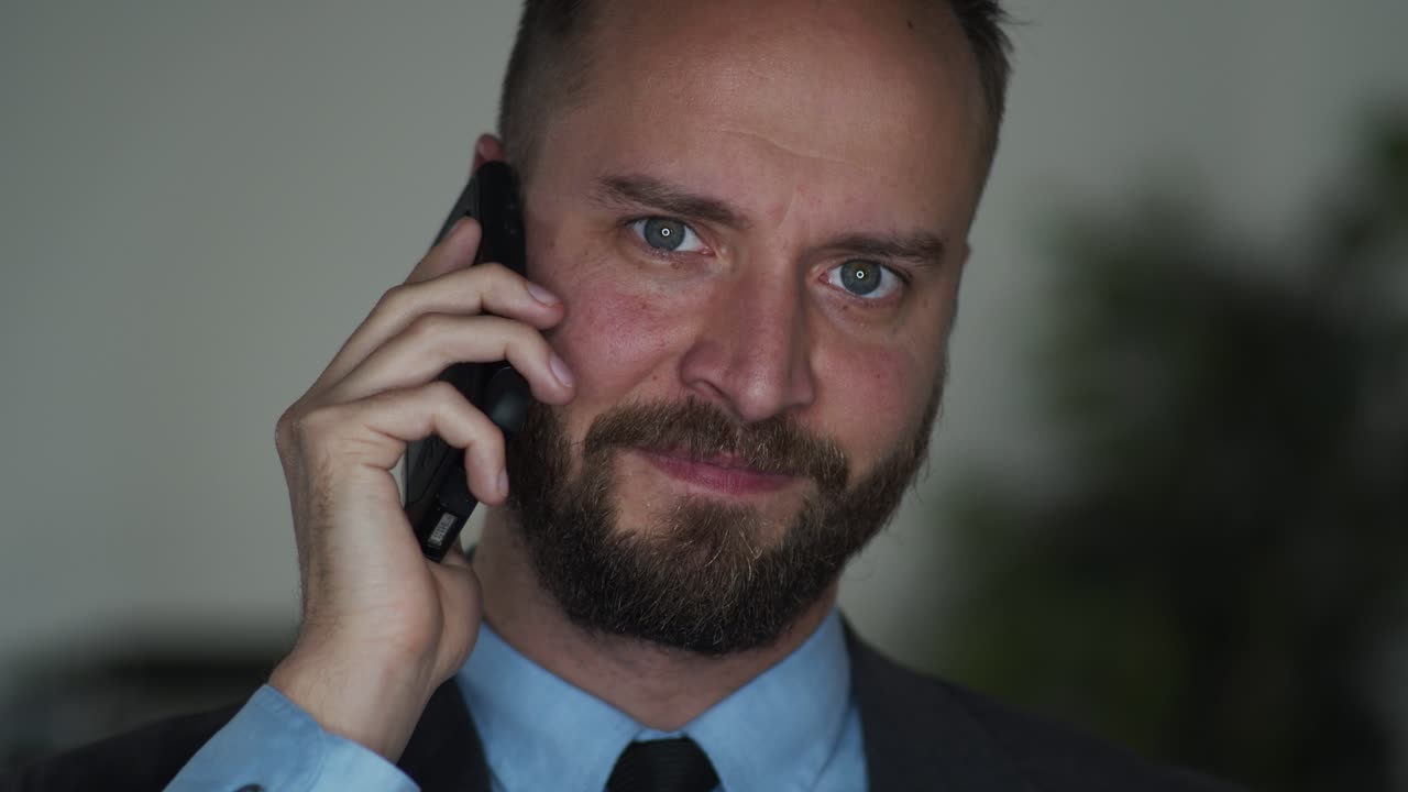 Bearded businessman on cellphone, talks and looks in the camera and smiles