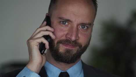 Bearded-businessman-on-cellphone,-talks-and-looks-in-the-camera-and-smiles