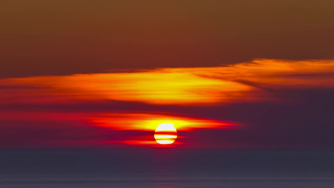 Bredt horisontalt udsyn til en glødende gylden sol nær den øverste tredjedel af billedrammen over en rolig havhorisont. Varmt orange og gult lys oplyser lagdelte lave skyer.
