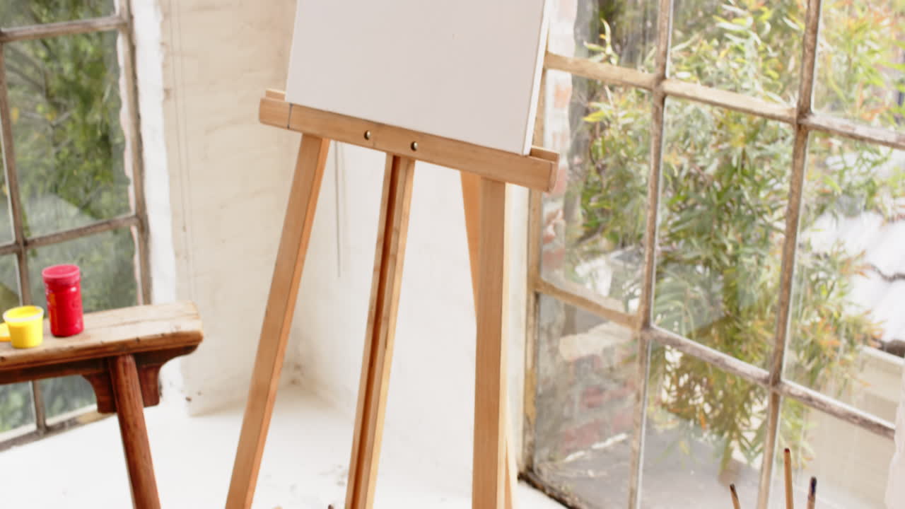 Easel with colorful abstract painting near window in cozy art studio, at home