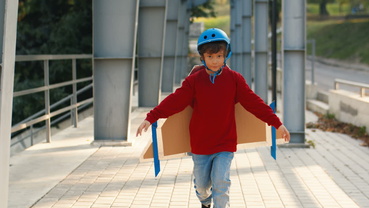 Cute Little Boy In Helmet And Red Sweater With Cardboard Airplane Wings Running On A Bridge On A Sunny Day