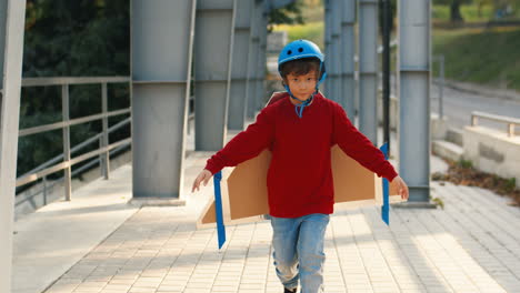 lindo niño pequeño con casco y suéter rojo con alas de avión de cartón corriendo en un puente en un día soleado