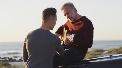 happy caucasian gay male couple sitting on car playing guitar and talking at the beach