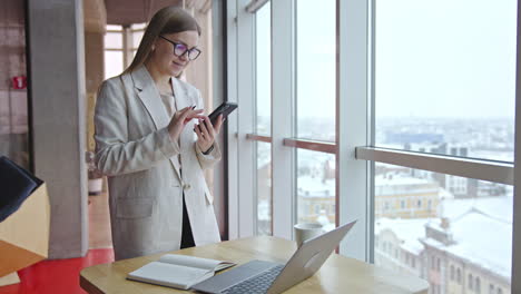 woman working on a laptop and smartphone in a modern office near a window.